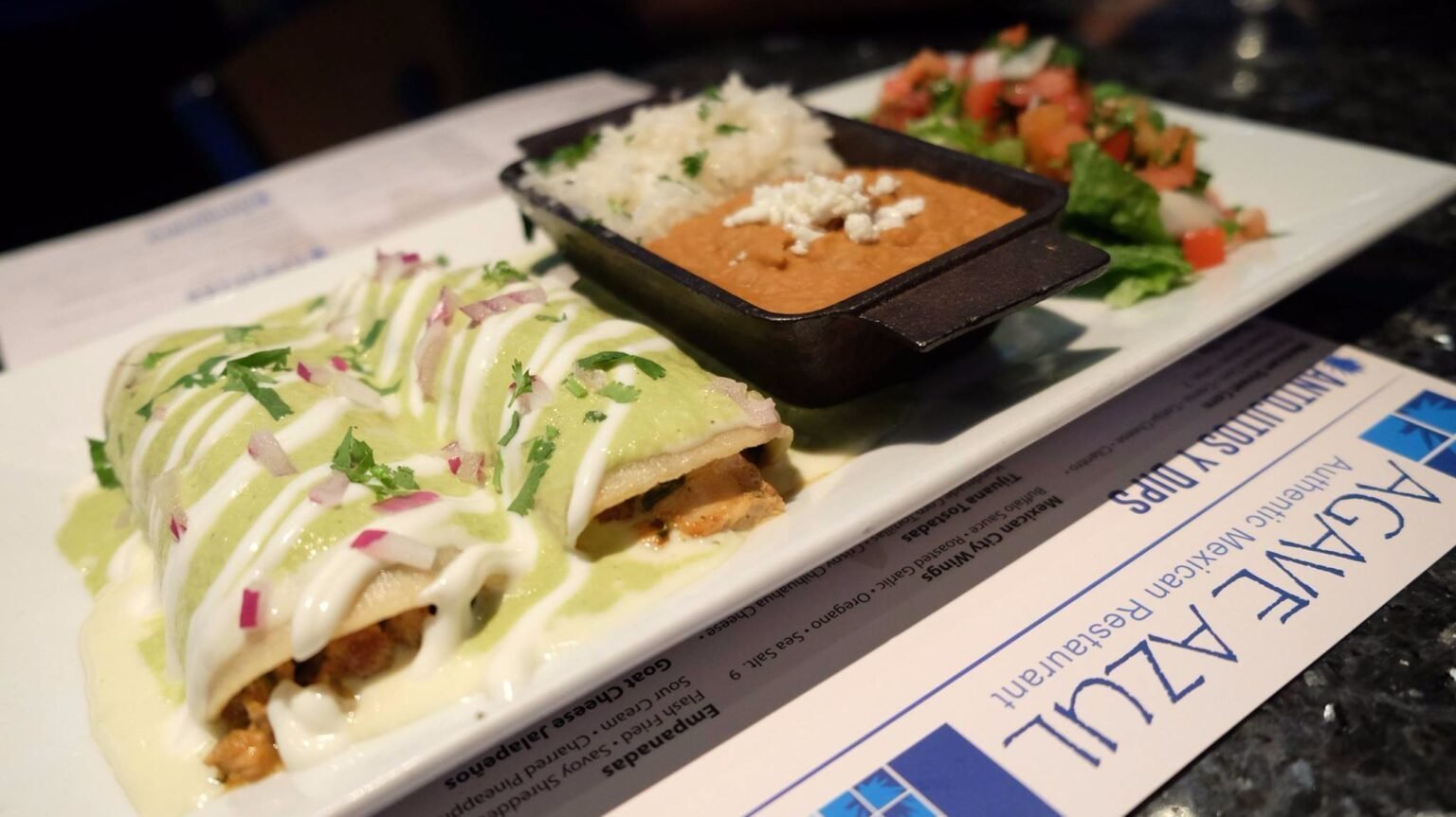 Enchiladas topped with green sauce and sour cream, served with rice, refried beans, and a fresh salad, featuring Agave Azul branding in the background.