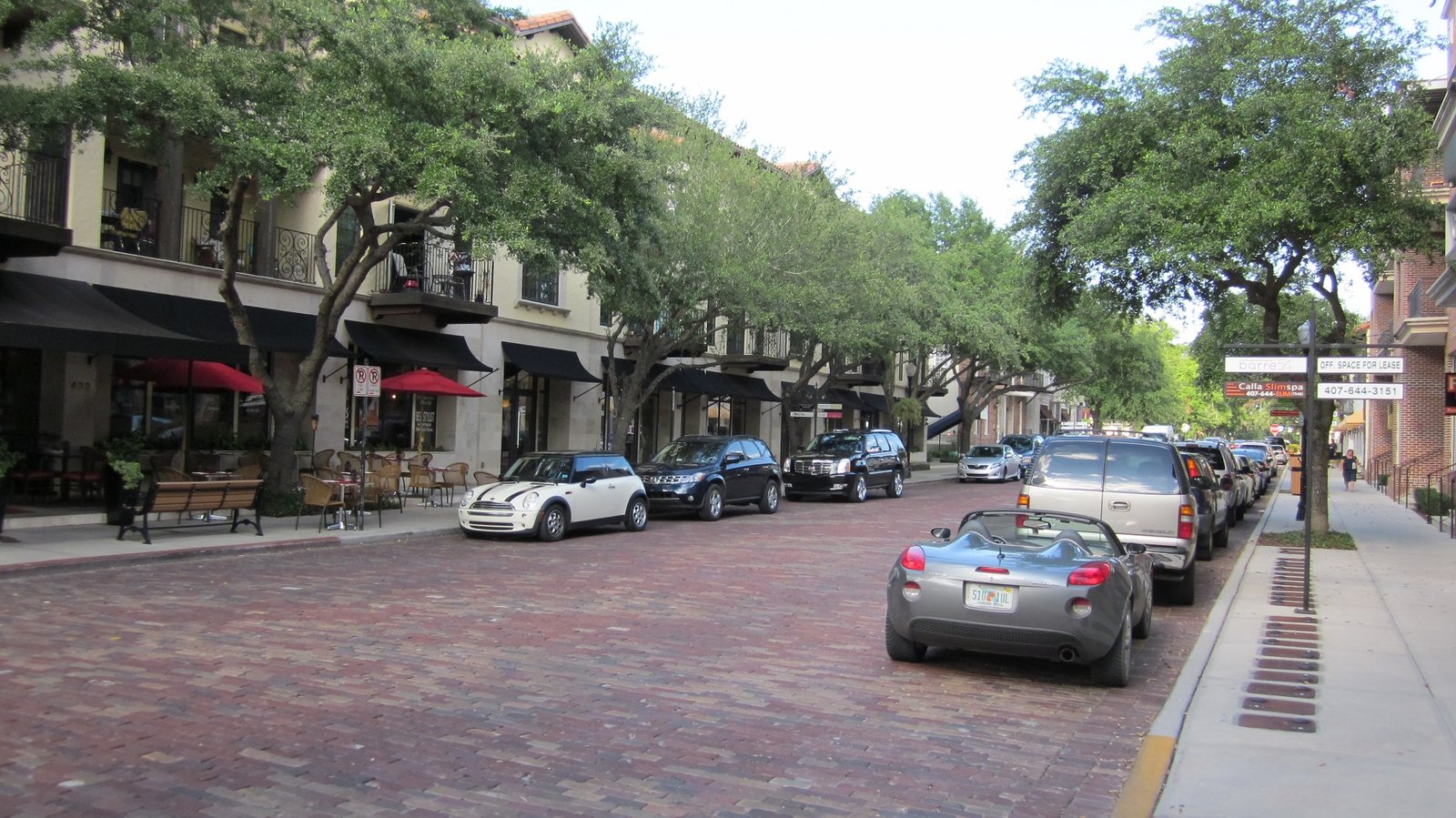 Street view of Park Avenue in Winter Park, Florida, featuring parked cars, outdoor dining areas with red umbrellas, and storefronts, highlighting the city's shopping and dining district.
