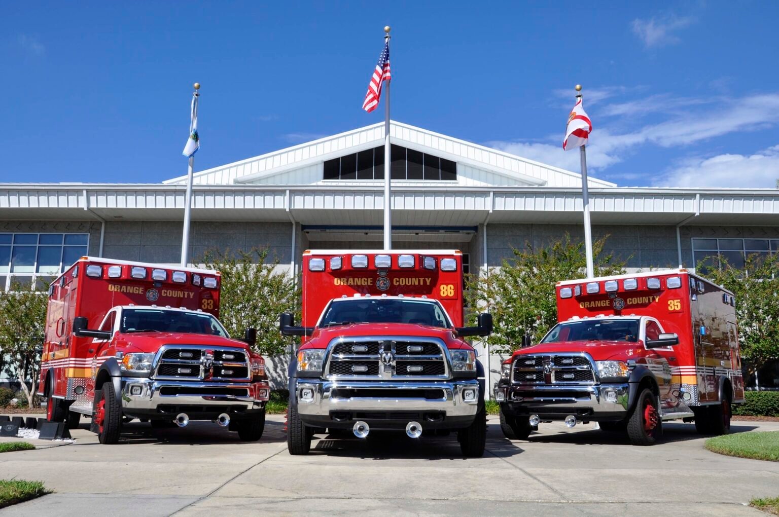 Three red ambulances from Orange County parked in front of a modern building, with flags flying above, representing emergency services in Windermere, Florida.