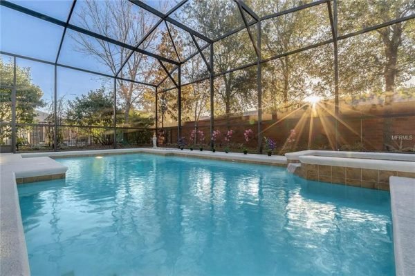 Screened pool area with clear blue water, surrounded by lush greenery and sunlight streaming through trees, showcasing a serene outdoor space in Windermere, FL.