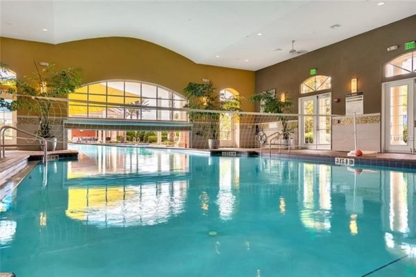 Indoor swimming pool in Trilogy over 55 community, featuring clear water, volleyball net, lush greenery, and large windows reflecting natural light.