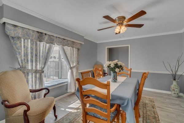 Dining room featuring a wooden table with blue tablecloth, surrounded by chairs, floral centerpiece, and a cozy armchair, with large windows draped in patterned curtains and a ceiling fan overhead, reflecting the inviting atmosphere of a Windermere home.