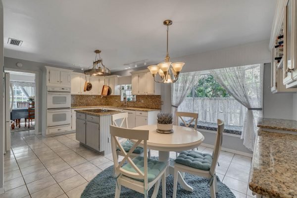 Bright, modern kitchen featuring white cabinetry, granite countertops, and a cozy dining area with a round table and teal accents, showcasing large windows with natural light and a view of the outdoors.
