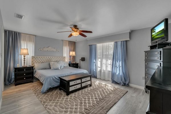 Master bedroom in Windermere, featuring a king-size bed with blue bedding, two bedside lamps, large windows with sheer curtains, a ceiling fan, and a dark wood dresser with a TV.