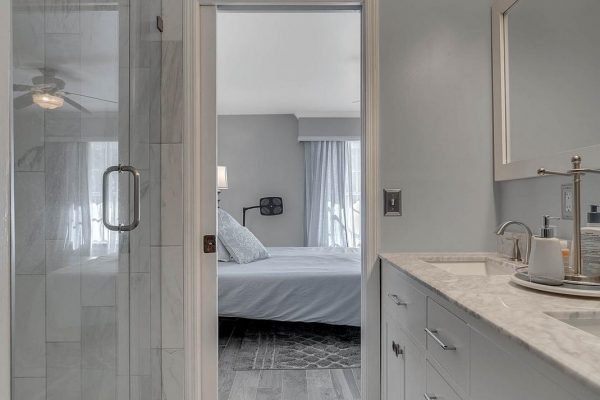 Master bathroom view leading to a well-lit bedroom, featuring modern fixtures, marble shower, and elegant vanity with natural light illuminating the space.