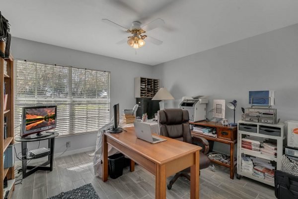 Home office space with wooden desk, laptop, and printer, featuring a window with blinds, shelving, and a television, reflecting a functional workspace in Windermere real estate listing.