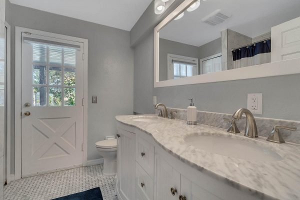 Modern bathroom featuring dual sinks with marble countertop, light gray walls, and a door leading outside, emphasizing spaciousness and natural light.