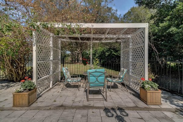 Outdoor seating area with a white pergola, featuring turquoise chairs and flower planters, surrounded by greenery in the backyard of a Windermere estate.