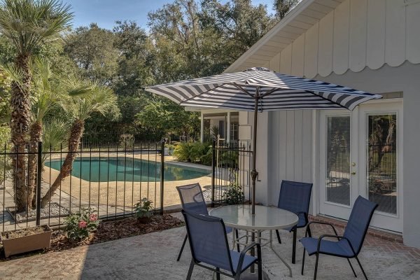 Backyard patio at 1421 Crescent Lake Dr, Windermere, FL, featuring a round table with four chairs, a striped umbrella, and a fenced swimming pool surrounded by lush greenery.