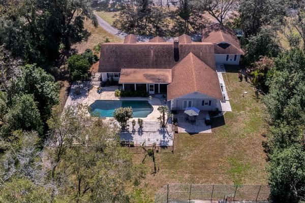 Aerial view of a spacious one-story estate at 1421 Crescent Lake Dr, Windermere, FL, featuring a large swimming pool, extensive decking, and a private tennis court, surrounded by mature trees.