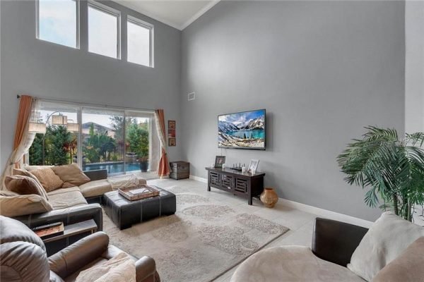 Spacious living room featuring high ceilings, large windows with views of the backyard pool, modern furnishings, and a mounted television, showcasing the elegant interior of a home in Winter Garden, FL.