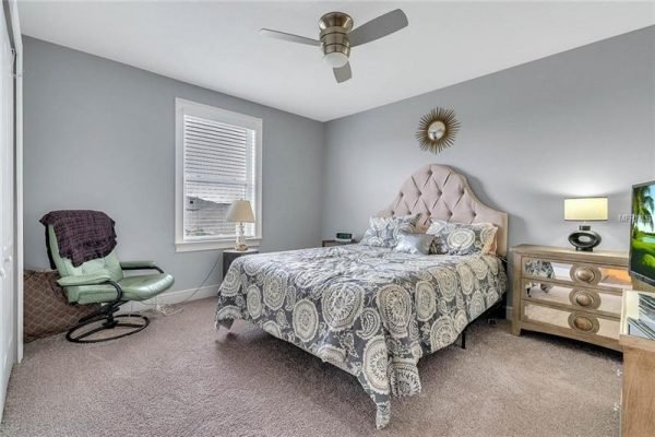 Bedroom featuring a queen-sized bed with decorative bedding, a modern ceiling fan, a cozy green chair, and a stylish nightstand, set in a well-lit space with gray walls and a window allowing natural light.