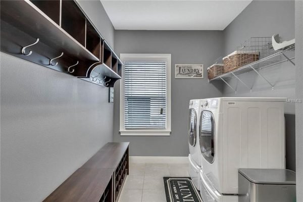 Laundry room featuring white washer and dryer, gray walls, wood shelving with hooks, and a window with blinds, emphasizing functionality and organization.