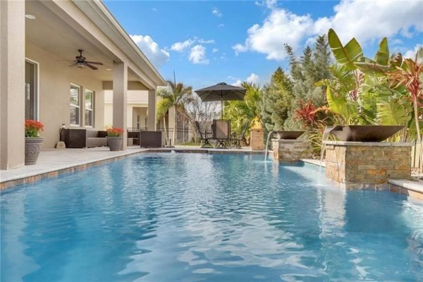 Poolside view of a luxury home in Winter Garden, featuring a heated saltwater pool, tropical landscaping, and covered seating area, showcasing outdoor entertainment space.