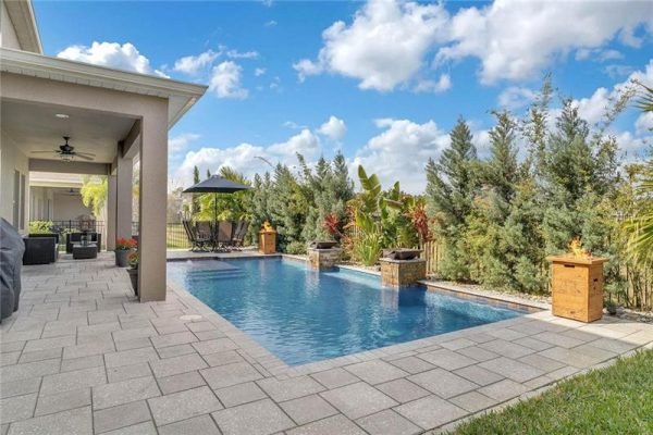 Backyard view of a luxury home in Winter Garden, featuring a heated saltwater pool, tropical landscaping, and a covered seating area, ideal for entertaining.