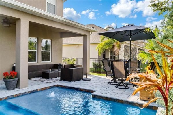Backyard view of a home in Winter Garden, featuring a heated saltwater pool, outdoor seating area with an umbrella, and tropical landscaping, highlighting the property's entertaining space and premium lot in the Reserve of Carriage Pointe community.