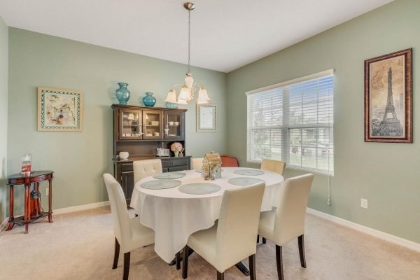 Dining room with a round table set for six, beige chairs, green walls, and a decorative hutch, featuring natural light from a window and an Eiffel Tower print on the wall, highlighting the spacious layout of the home at 15522 Sandfield Loop, Winter Garden, FL.