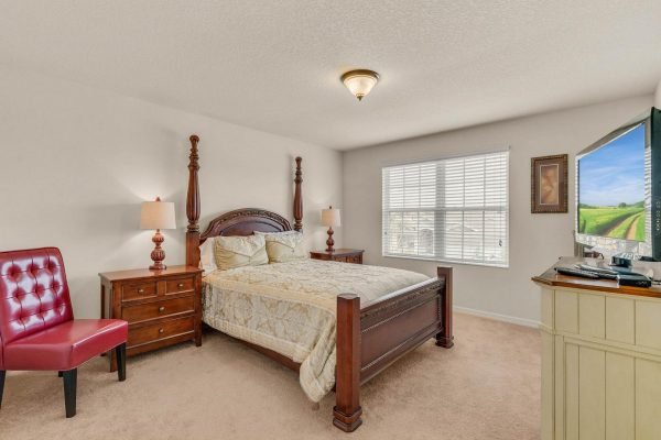 Cozy bedroom featuring a wooden bed frame with decorative posts, complemented by a red accent chair, bedside lamps, and a TV, showcasing natural light from a nearby window in a well-maintained home in Winter Garden, FL.