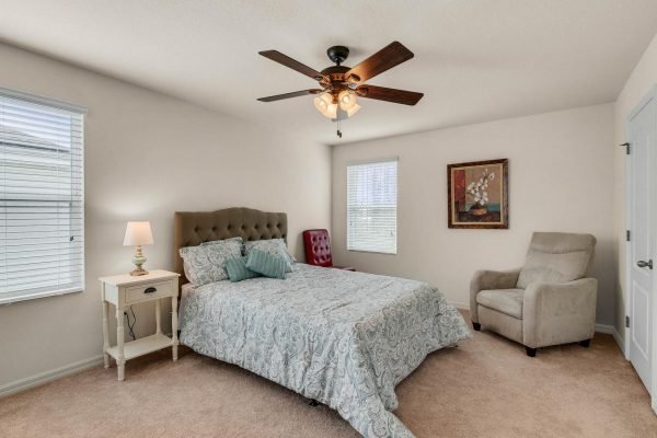 Cozy bedroom featuring a queen-sized bed with a tufted headboard, bedside lamp, and accent chair, complemented by natural light from windows and decorative artwork, highlighting the inviting atmosphere of the home at 15522 Sandfield Loop, Winter Garden, FL.