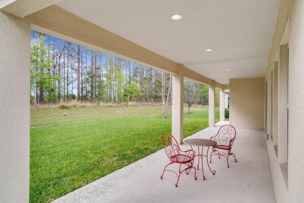 Covered lanai with red wrought iron table and chairs overlooking lush green lawn and trees, showcasing outdoor living space at 15522 Sandfield Loop, Winter Garden, FL.