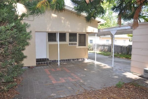 Patio area of 1006 Venetian Ave, Orlando, FL, featuring brick pavers, fenced yard, and adjacent carport, surrounded by mature trees.