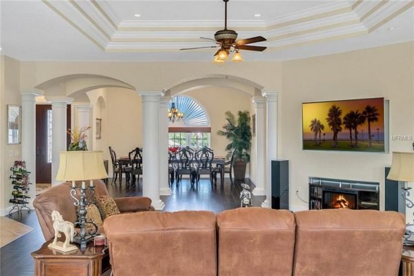 Elegant living room interior featuring a cozy brown sofa, decorative lamp, and detailed ceiling design, with a view into a dining area and a large window showcasing outdoor greenery, complemented by a wall-mounted TV displaying a serene landscape.