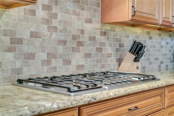 Modern kitchen featuring a Bosch gas range with a sleek granite countertop, stylish backsplash of mixed stone tiles, and a knife block, showcasing elegant design elements in the luxury property at 11114 Romance Ct, Winter Garden, FL.