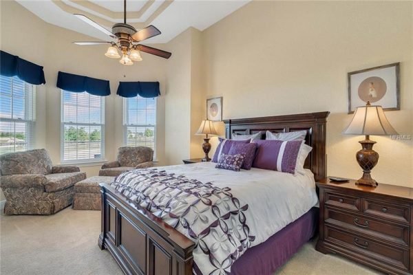 Master bedroom in Winter Garden, Florida featuring a king-sized bed with purple accents, two armchairs, large windows with blue valances, and a ceiling fan, showcasing elegance and spaciousness in a luxury home.