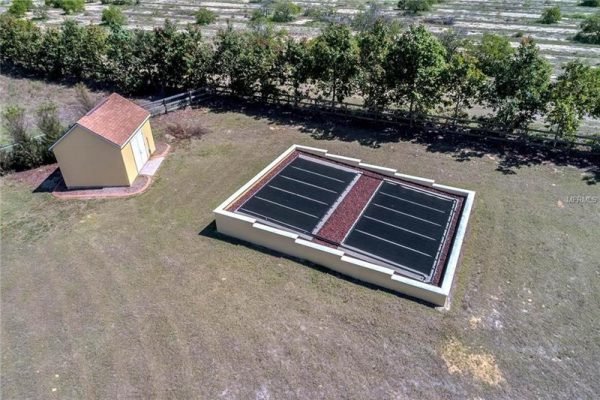 Aerial view of a property in Winter Garden, Florida, featuring a large solar panel installation and a small yellow shed on a spacious grassy lot surrounded by trees.