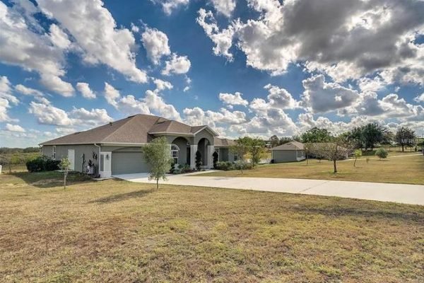 Exterior view of a large single-family home at 11630 Flynn Ct, Winter Garden, FL, showcasing a spacious front yard, two-car garage, and a scenic landscape with trees under a partly cloudy sky.