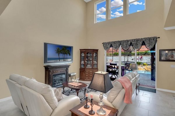 Bright family room featuring white leather sofas, a wooden entertainment center with a beach scene TV, and large windows overlooking a private backyard with a pool, emphasizing the spacious and inviting atmosphere of the Hillsdale floor plan home at 15390 Sandfield Loop, Winter Garden, FL.
