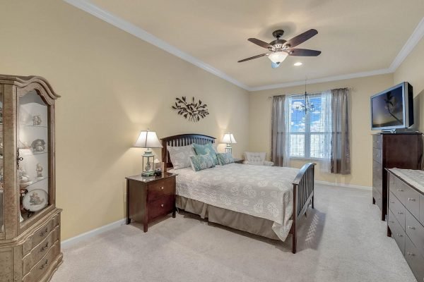 Master bedroom featuring a king-sized bed with decorative bedding, two bedside lamps, a dresser, and a glass display cabinet; natural light from window with sheer curtains, and ceiling fan, showcasing the spacious layout and neutral color palette of the home at 15390 Sandfield Loop, Winter Garden, FL.