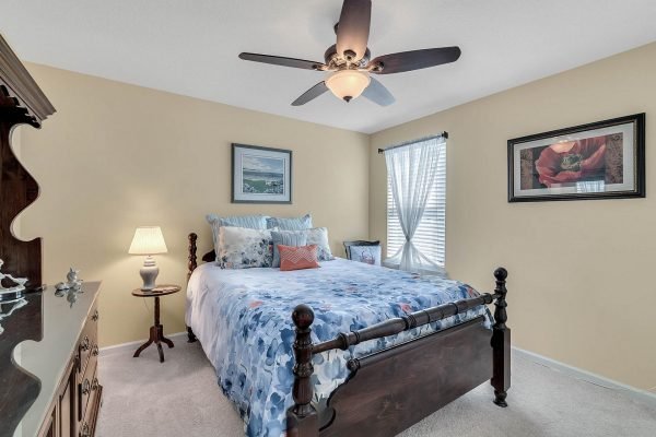 Cozy bedroom featuring a queen-sized bed with blue and white bedding, decorative pillows, a bedside lamp, and a ceiling fan, complemented by light yellow walls and framed artwork.