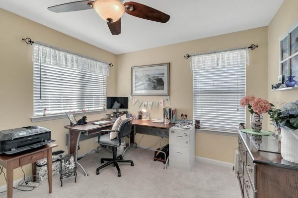 Home office with desk and computer, natural light from windows, light yellow walls, decorative plants, and ceiling fan, showcasing a cozy workspace in the property at 15390 Sandfield Loop, Winter Garden, FL.