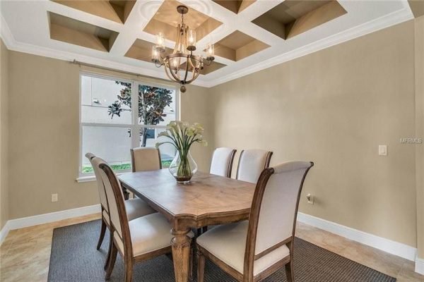 Dining room with coffered ceiling, modern chandelier, and elegant wooden table set for six, featuring a vase of flowers and natural light from large window, showcasing upscale interior design in Windermere, FL home.