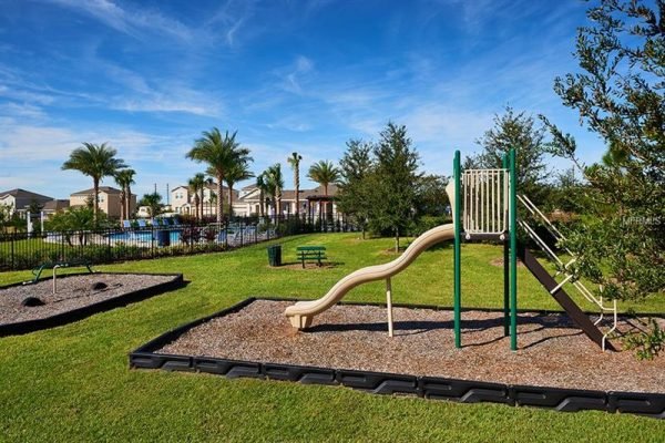 Playground area with slide and swings, surrounded by lush grass and palm trees, featuring a community pool in the background, highlighting amenities in the Hilltop community of Apopka, FL.