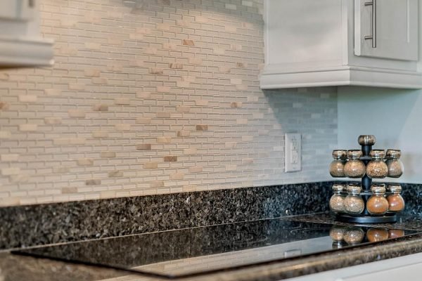Modern kitchen featuring a sleek black granite countertop, decorative backsplash with textured tiles, and a spice rack with various jars, reflecting the upscale design of the Windermere estate at 11637 Brickyard Pond Lane.