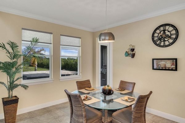 Dining area featuring a glass table set for four, surrounded by wicker chairs, with a palm plant in a pot, large windows providing natural light, and a wall clock, highlighting the spacious and inviting ambiance of the Windermere lakefront estate.