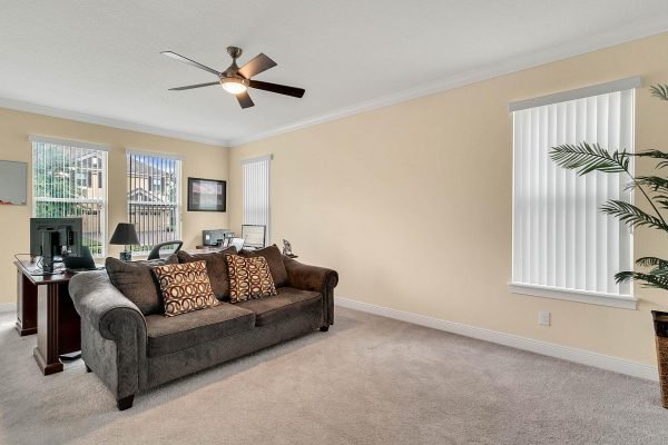 Cozy living room featuring a plush sofa with decorative pillows, a ceiling fan, and a workspace with a desk and lamp, brightened by natural light from large windows with vertical blinds.