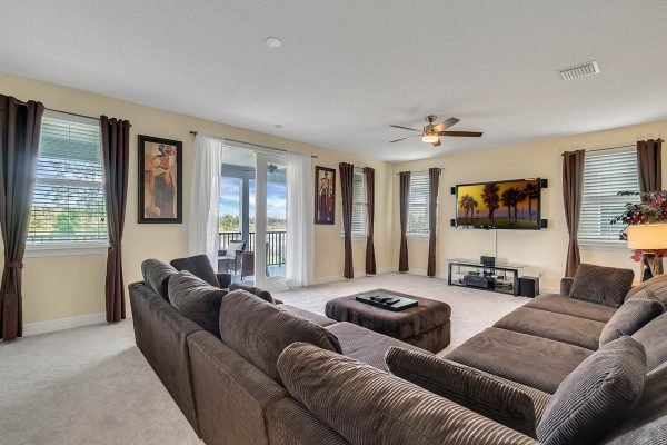 Spacious living room featuring a large sectional sofa, decorative artwork, and a wall-mounted TV, with natural light from multiple windows and sliding doors leading to a balcony, ideal for a lakefront estate in Windermere, FL.