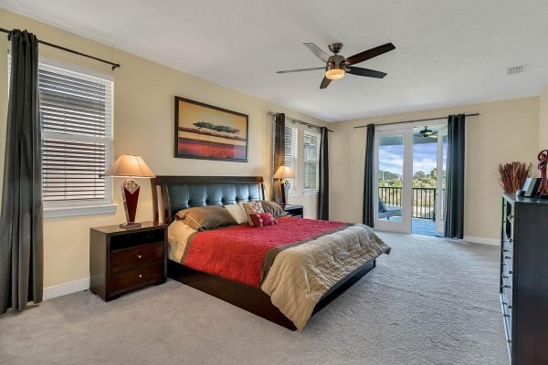 Master bedroom in Windermere estate featuring king-size bed with red and beige bedding, nightstands with lamps, large windows, and double doors leading to a balcony with scenic views.