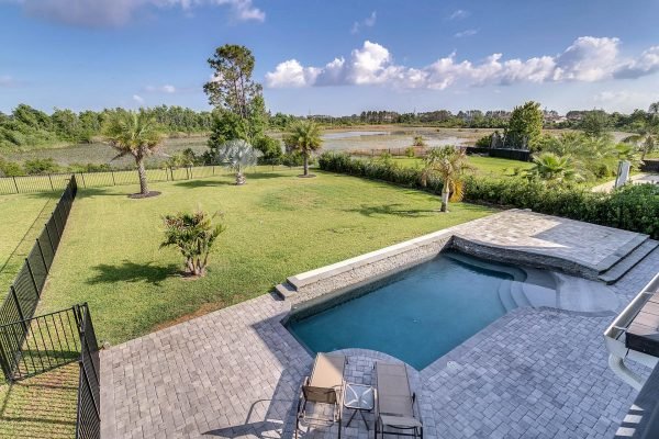 Lakefront backyard view of 11637 Brickyard Pond Lane, featuring a swimming pool, paver patio, and landscaped yard with palm trees, overlooking Lake Burden in Windermere, FL.