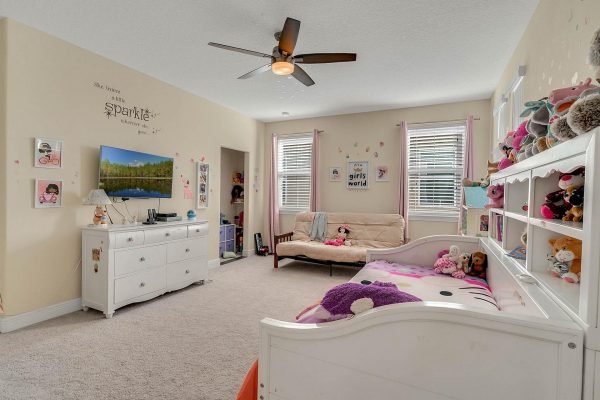 Spacious children's bedroom with white furniture, pink accents, and a cozy seating area, featuring a TV, decorative wall art, and a collection of stuffed animals.