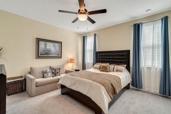 Cozy bedroom featuring a king-sized bed with a tufted headboard, a comfortable sofa, decorative pillows, and large windows with blue curtains, creating a bright and inviting atmosphere.