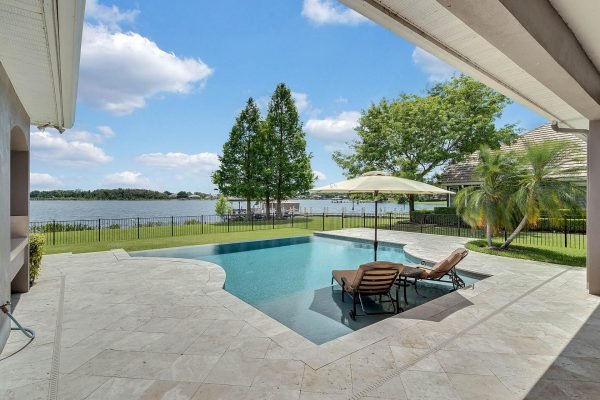 Private lakefront view from 2039 Water Key Drive, Windermere, featuring a solar heated infinity edge pool, travertine patio, and lush greenery.