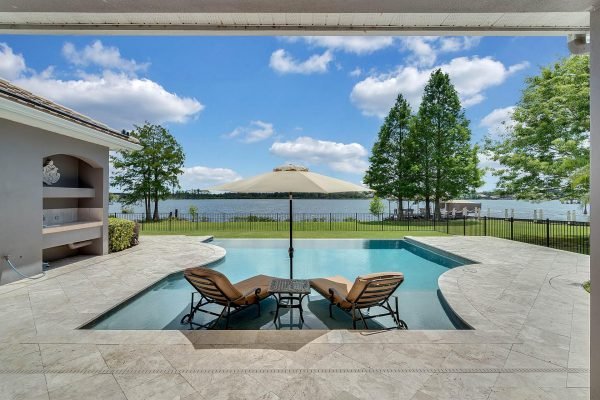 Lakefront view from a luxury home in Windermere, featuring a saltwater infinity pool, two lounge chairs under an umbrella, and expansive natural scenery with trees and water.