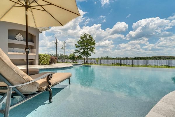 Lakefront infinity edge pool with lounge chair and umbrella, overlooking Lake Roberts in Windermere, Florida, featuring a clear blue sky and lush greenery.
