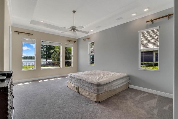 Spacious master bedroom with lake views, large windows, and ceiling fan, featuring an unmade bed and plush carpet, located in a private lakefront home in Windermere, FL.