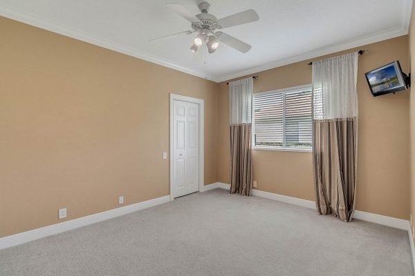 Empty bedroom with beige walls, ceiling fan, and window dressed with light curtains, showcasing a clean, spacious layout.