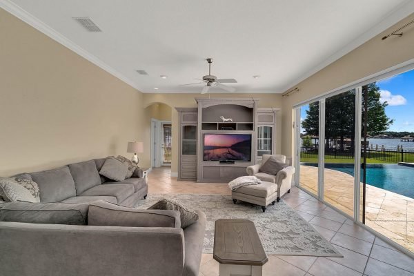 Living room featuring a gray sectional sofa, armchair, and entertainment center with a large TV, showcasing views of a lake and backyard pool through sliding glass doors, in a private lakefront home at 2039 Water Key Drive, Windermere, FL.